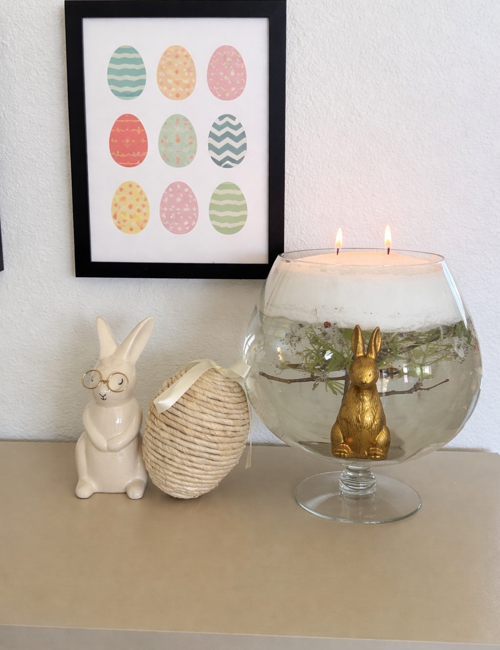 Spring home decor vignette with framed pastel Easter egg artwork, ceramic bunny figurines, and a glass bowl candle featuring floating greenery and a gold rabbit centerpiece on a neutral tabletop.