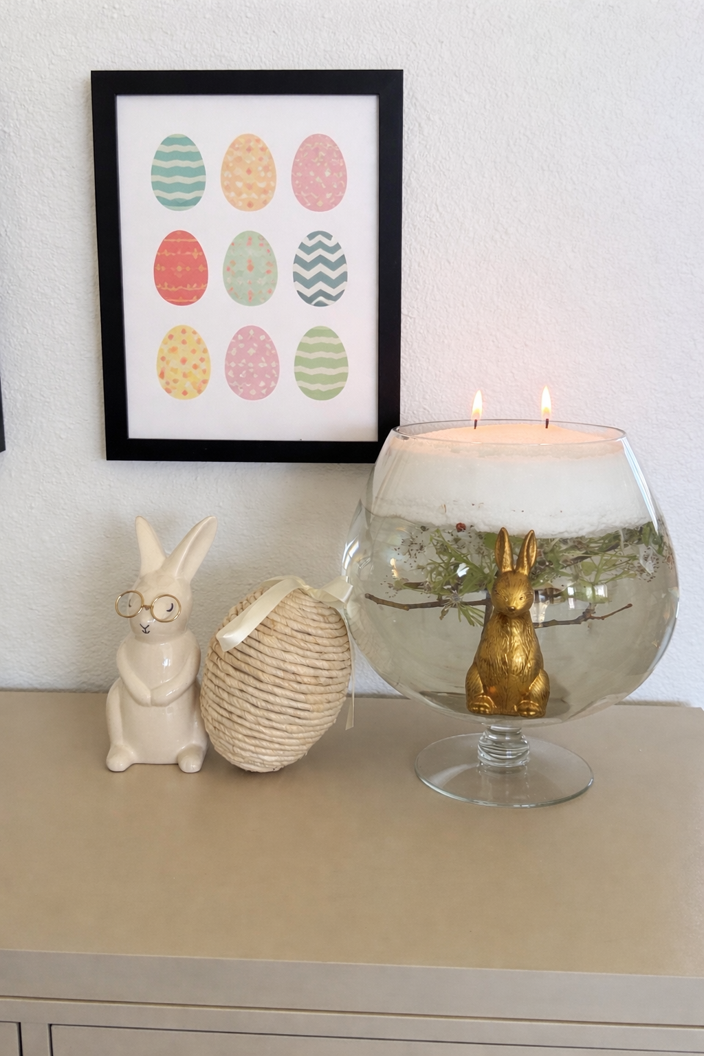 Spring home decor vignette with framed pastel Easter egg artwork, ceramic bunny figurines, and a glass bowl candle featuring floating greenery and a gold rabbit centerpiece on a neutral tabletop.