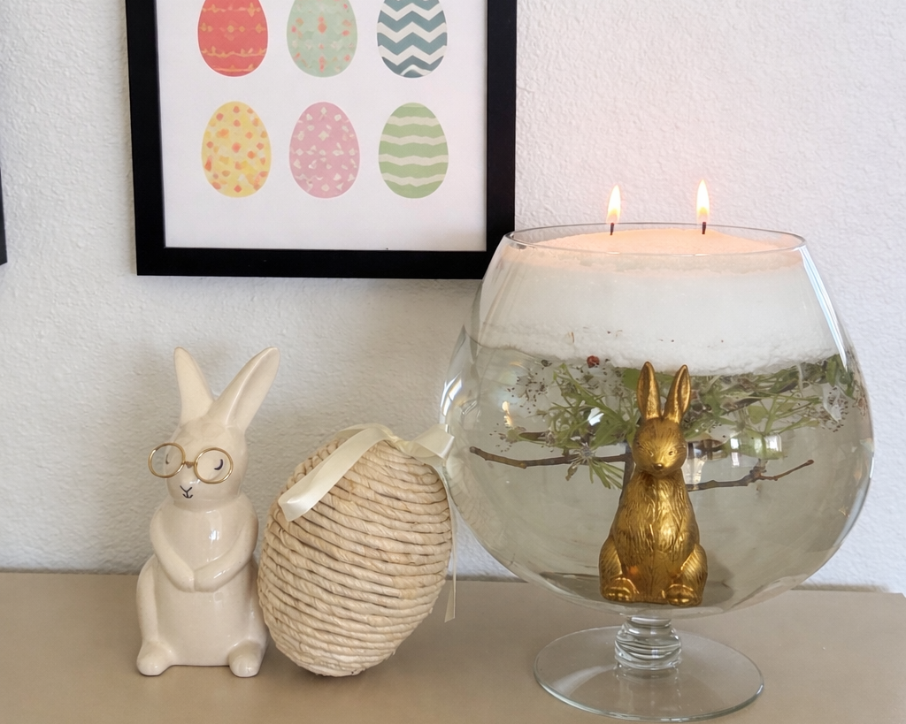 Spring home decor vignette with framed pastel Easter egg artwork, ceramic bunny figurines, and a glass bowl candle featuring floating greenery and a gold rabbit centerpiece on a neutral tabletop.